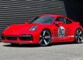 A red Porsche 911 Sport Classic with a "60" racing decal and "Porsche" branding on the side, parked on pavement in front of a dark striped metal wall—an homage to Porsche history.