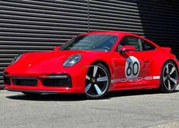 A red Porsche 911 Sport Classic with a "60" racing decal and "Porsche" branding on the side, parked on pavement in front of a dark striped metal wall—an homage to Porsche history.