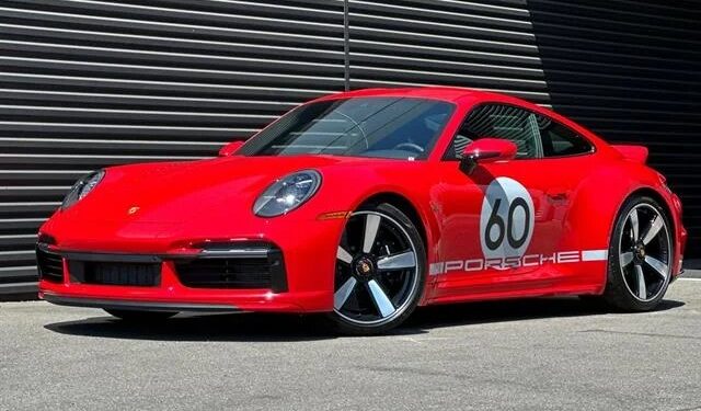 A red Porsche 911 Sport Classic with a "60" racing decal and "Porsche" branding on the side, parked on pavement in front of a dark striped metal wall—an homage to Porsche history.