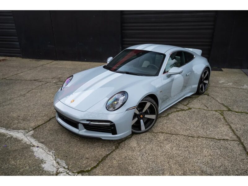 A silver Porsche 911 Sport Classic is parked on a cracked concrete surface in front of a black building with large garage doors, capturing a piece of Porsche history in every detail.