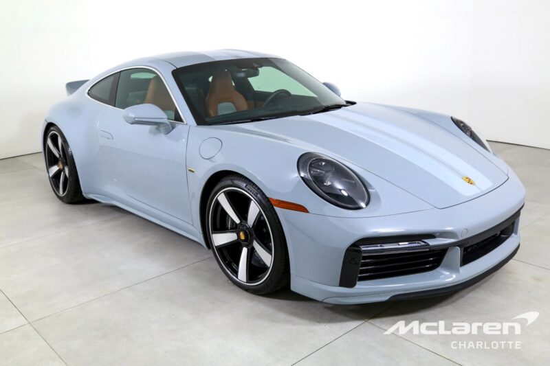 A light blue Porsche 911 Sport Classic coupe is parked indoors on a light-colored floor, with "McLaren Charlotte" branding visible in the bottom right corner.