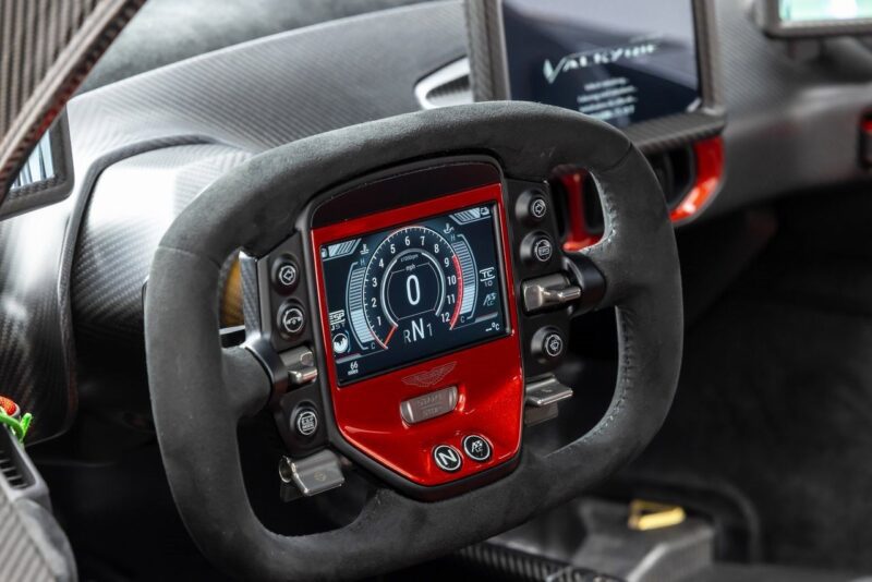 Close-up of a racing-style steering wheel with digital display and controls inside an ultra-rare Aston Martin Valkyrie, featuring the iconic logo and carbon fiber interior elements, offered by O’Gara Coach.