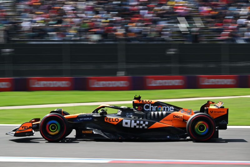 A McLaren Formula 1 car in striking orange and black races on a track, with blurred grandstands and spectators in the background.