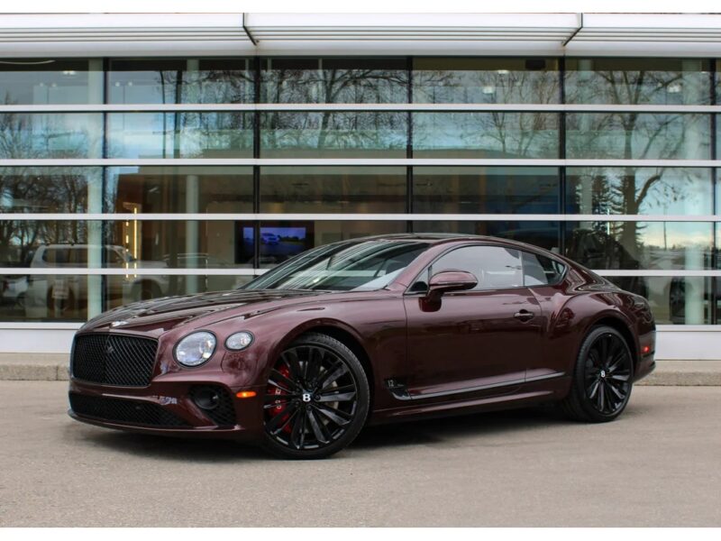 A maroon Bentley Continental GT, the epitome of a luxury tourer, is parked in front of a modern glass building.