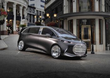 A futuristic silver Mercedes-Benz Vision V concept minivan with large wheels is parked on a cobblestone street in an upscale urban shopping area.
