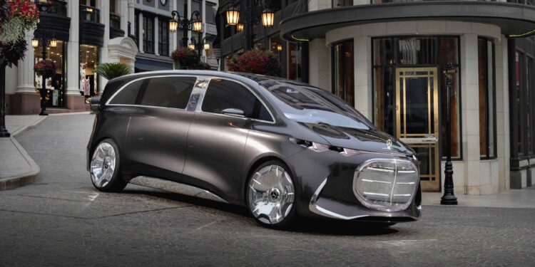A futuristic silver Mercedes-Benz Vision V concept minivan with large wheels is parked on a cobblestone street in an upscale urban shopping area.