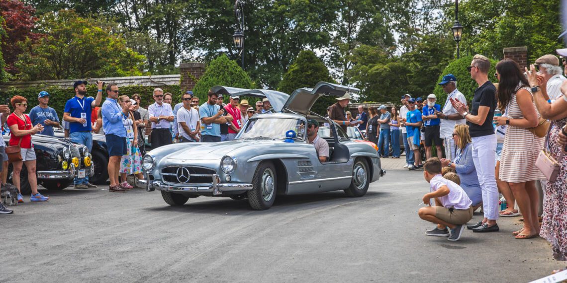 A silver vintage Mercedes-Benz with gullwing doors open drives on a road, surrounded by a crowd of people at the Misselwood Concours d’Elegance, celebrating its 15th Anniversary with record attendance and eager photographers.