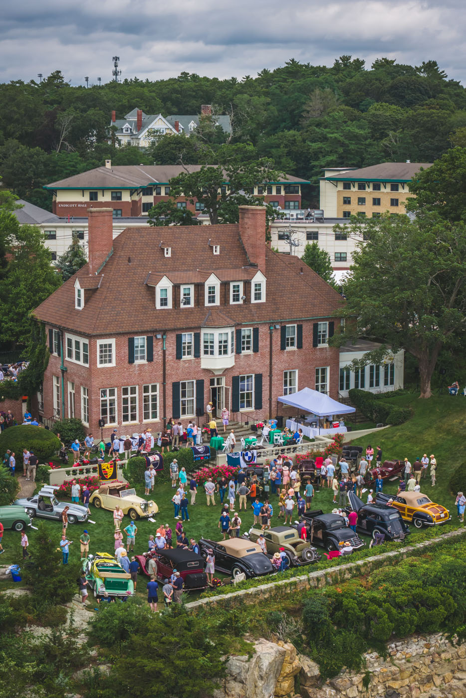 A large crowd gathers on the lawn of a brick mansion for the Misselwood Concours d’Elegance 15th Anniversary, viewing a stunning display of classic cars with buildings and trees in the background under a cloudy sky.