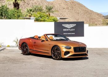 A gold Bentley Continental GTC convertible is parked in front of a Bentley dealership sign, with mountains and palm trees in the background, showcasing this iconic luxury tourer.