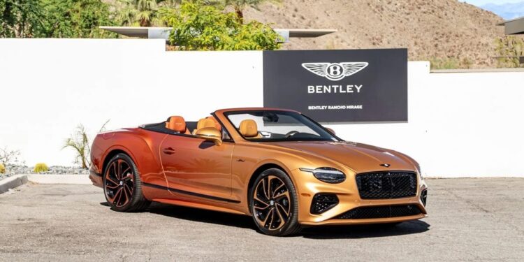 A gold Bentley Continental GTC convertible is parked in front of a Bentley dealership sign, with mountains and palm trees in the background, showcasing this iconic luxury tourer.