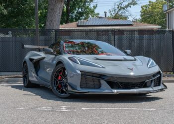 A gray 2025 Corvette ZR1 sports car is parked on a street in front of a black metal fence and trees, seen in daylight.