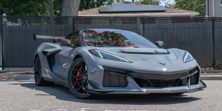 A gray 2025 Corvette ZR1 sports car is parked on a street in front of a black metal fence and trees, seen in daylight.