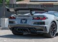 A grey Corvette ZR1 with a large rear wing and quad exhaust pipes is parked on a street near a black fence, available now from Status Auto Group.