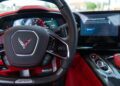 Close-up of a Corvette ZR1 C8 steering wheel with digital dashboard, control buttons, and an upgrade prompt on screen. The 2025 Corvette interior features striking red and carbon fiber accents.