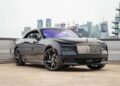 A black Rolls-Royce Spectre electric super coupe with a prominent grille is parked on a rooftop, set against a city skyline and construction cranes in the background.