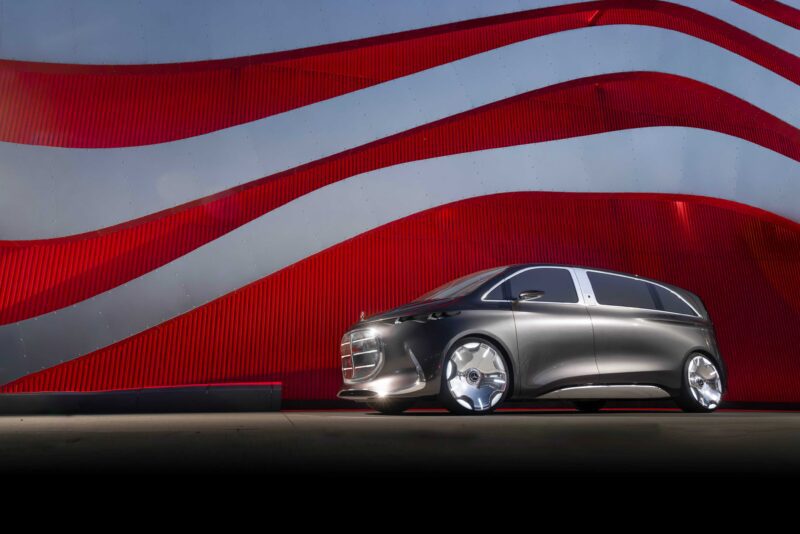 A sleek, silver Mercedes-Benz Vision V concept minivan is parked in front of a modern building with red and silver wavy panels.
