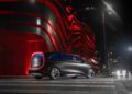 A futuristic Mercedes-Benz Vision V van luxury concept with illuminated accents is parked on a crosswalk at night, set against a modern building adorned with red illuminated wave patterns in the background.
