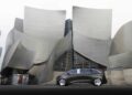 A futuristic silver Mercedes-Benz Vision V concept luxury van is parked in front of a modern building with curved, metallic walls and glass windows.