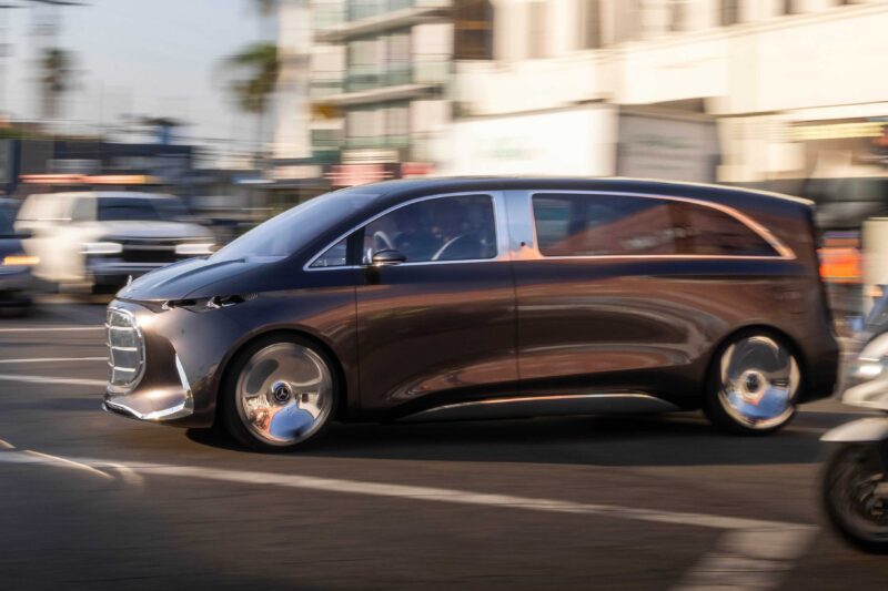 A modern, dark-colored Mercedes-Benz Vision V minivan with a sleek design and van luxury drives through a busy city street during the day.