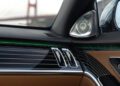 Close-up of a Mercedes-Maybach S 680’s luxurious dashboard and door panel featuring leather, wood trim, metallic air vents, a speaker, and green ambient lighting—part of the limited edition Emerald Isle visible beyond the window.