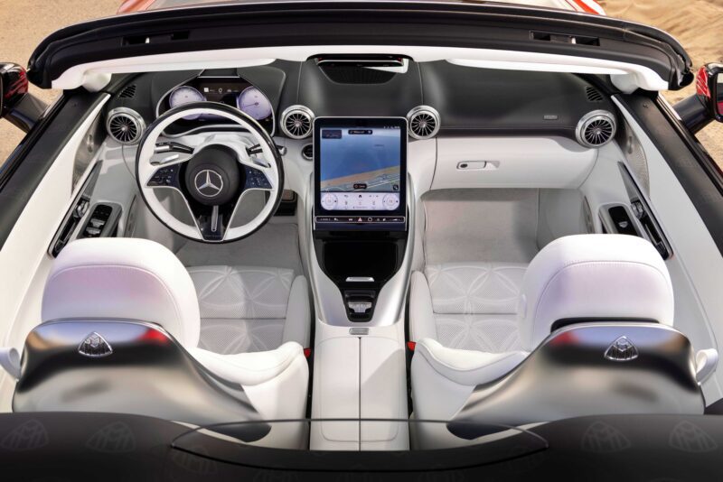 View of an ultra-luxury roadster interior, showcasing a Mercedes-Maybach SL 680 steering wheel, large central touchscreen, and two white leather seats adorned with Maybach Monogram Series logos.