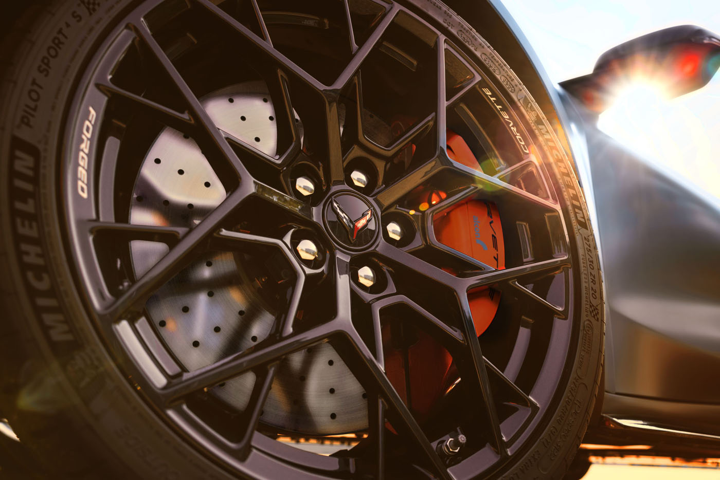 Close-up of a black alloy wheel with a Corvette logo, Michelin Pilot Sport tire, and red brake caliper on the 2026 Corvette ZR1X Quail Silver Limited Edition, sun flare accenting its sporty style at Monterey Car Week.