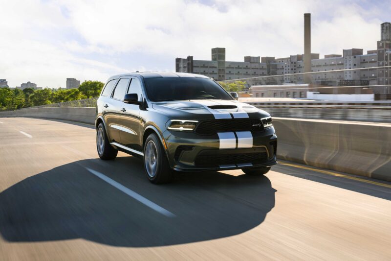 A black 2026 Dodge Durango Hellcat Jailbreak with white racing stripes drives on a city highway, showcasing its 710 HP and hinting at the 6 million possible builds available, with buildings and trees in the background.