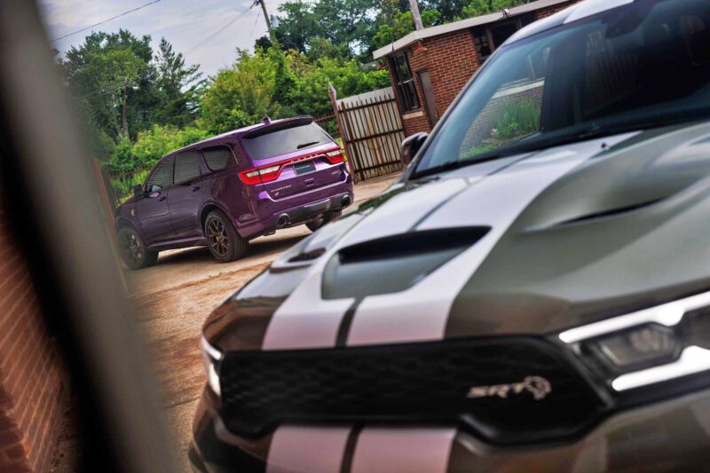 A purple 2026 Dodge Durango Hellcat Jailbreak, boasting 710 HP and endless build options, and a gray car with white racing stripes are parked outside near brick buildings on a cloudy day.