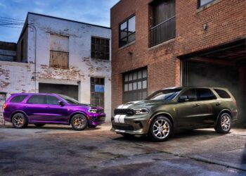 Two 2026 Dodge Durango Hellcat SUVs, one purple and one dark green with white stripes, are parked outside brick industrial buildings on a concrete lot, showcasing the bold presence of the Jailbreak edition.