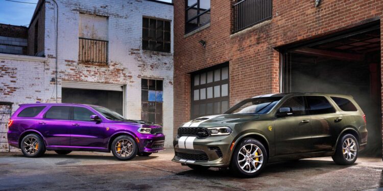 Two 2026 Dodge Durango Hellcat SUVs, one purple and one dark green with white stripes, are parked outside brick industrial buildings on a concrete lot, showcasing the bold presence of the Jailbreak edition.