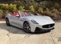 A silver Maserati convertible sports car with a red interior is parked on a concrete surface near bushes and a fence, showcasing true Italian luxury.