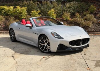 A silver Maserati convertible sports car with a red interior is parked on a concrete surface near bushes and a fence, showcasing true Italian luxury.