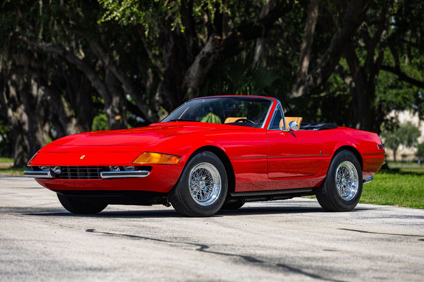 A classic red convertible sports car with chrome wheels is parked on a paved road with trees and grass in the background, ready to turn heads at the upcoming Mecum Auction during Monterey 2025.