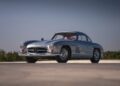 A classic silver Mercedes-Benz 300SL coupe with gullwing doors, one of the million-dollar cars, parked on a paved surface against a clear sky and distant greenery—an iconic presence perfect for the Monterey 2025 Auction by Mecum.