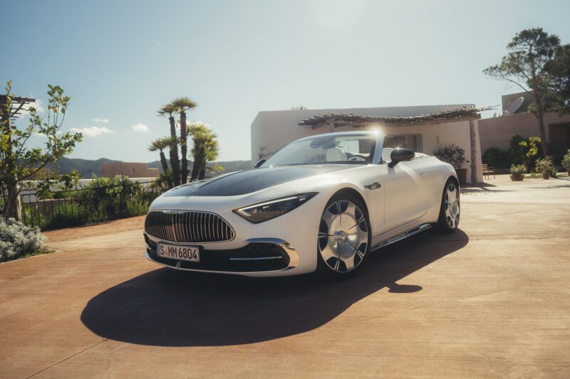 A silver and black Maybach SL 680 luxury convertible with a sporty soft-top is parked on a sunlit driveway, surrounded by greenery and modern buildings in the background.