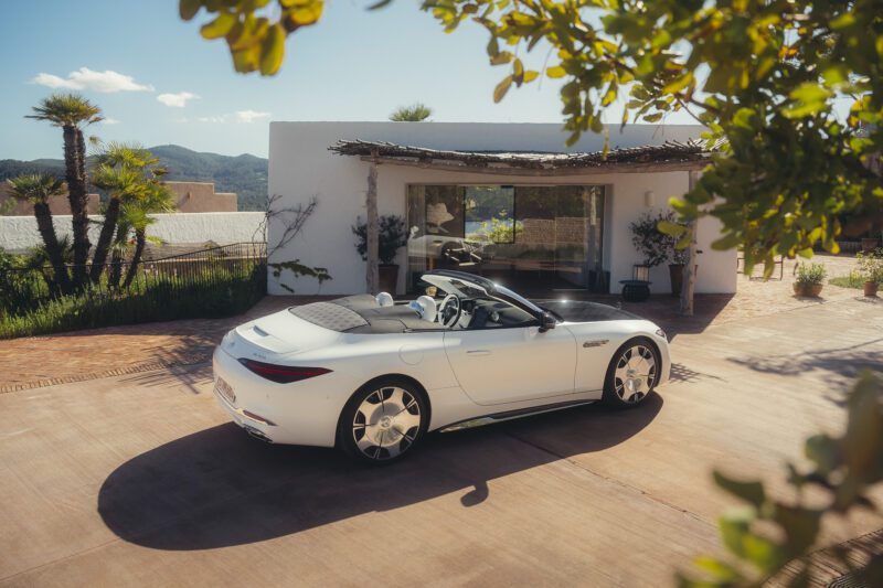 A sporty soft-top SL 680 Maybach convertible is parked in front of a modern house with a patio, lush plants, and large windows on a sunny day.