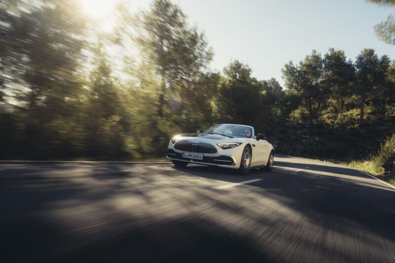 A sporty soft-top Maybach SL 680 convertible drives on a sunlit, tree-lined road, its sleek silhouette highlighted by the motion blur in the background.