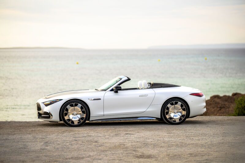 A white Maybach SL 680 convertible sports car with a sporty soft-top is parked on a road by the seaside, roof down, facing left, beneath a clear sky.
