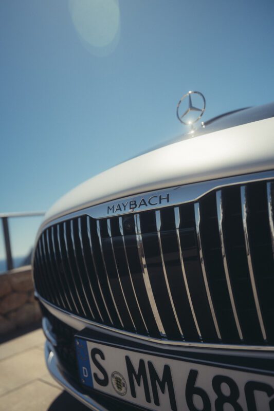 Close-up shot of the front grille and hood of a white Mercedes-Maybach car, showcasing the prominent Maybach badge and part of a license plate, with subtle sporty accents enhancing its luxurious appeal.