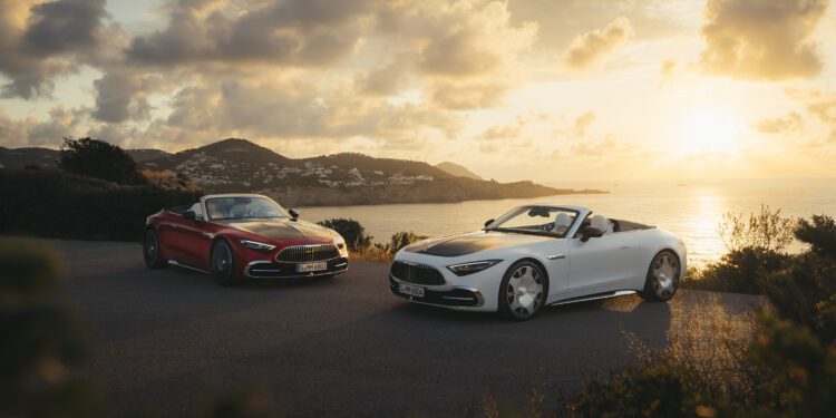 Two sporty soft-top convertibles, one red and one white, parked on a coastal road at sunset with the ocean and distant hills in the background, add a touch of Maybach luxury to the breathtaking scene.