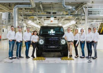 A group of ten people in white shirts and jeans stand around a black Mercedes-Benz G-Class SUV marked "600,000" inside an automotive factory.