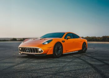 An orange CONCEPT AMG GT XX Mercedes-Benz sports car is parked on an empty, sunlit asphalt surface with clear skies, ready for an exhilarating drive around the world.