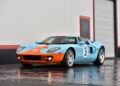 A blue and orange Ford GT sports car with number 3 decals is parked on a wet concrete surface in front of a closed garage door, reminiscent of the Million-Dollar Cars often seen at the Mecum Monterey 2025 Auction.