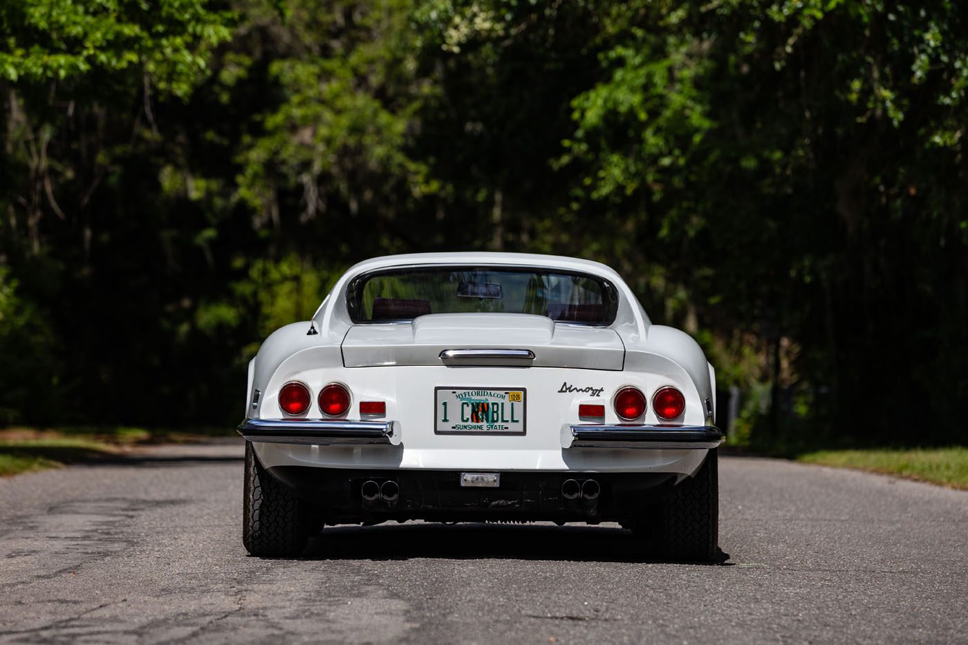 Rear view of a classic white sports car with round tail lights and a "1 CARBL" license plate, parked on a tree-lined road—an image reminiscent of Ferrari Legends at the Monterey auction or Mecum 2025.