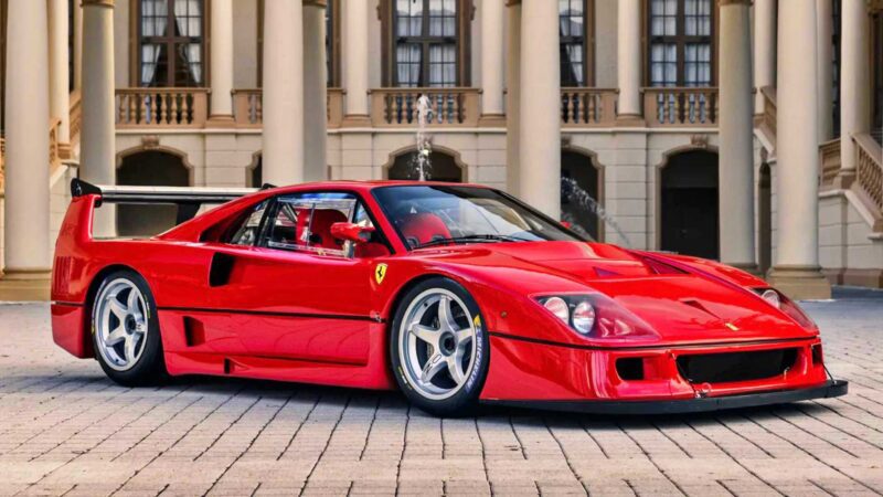 A red Ferrari F40 sports car, one of the 10 Most Expensive Cars To Sell At 2025 Monterey Car Week Auctions, is parked on a cobblestone driveway in front of a building with columns.