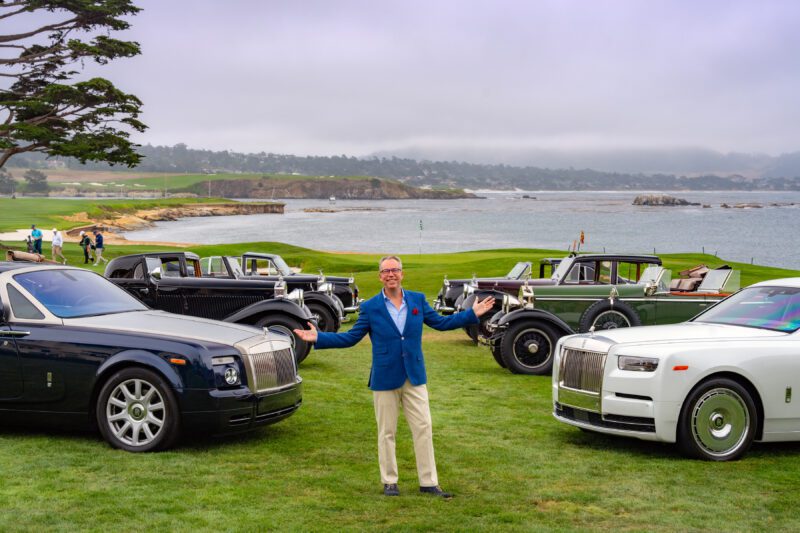 A man stands with arms outstretched on a grassy field at Pebble Beach, surrounded by vintage and modern Rolls-Royce Phantom cars, with a stunning coastal landscape in the background.