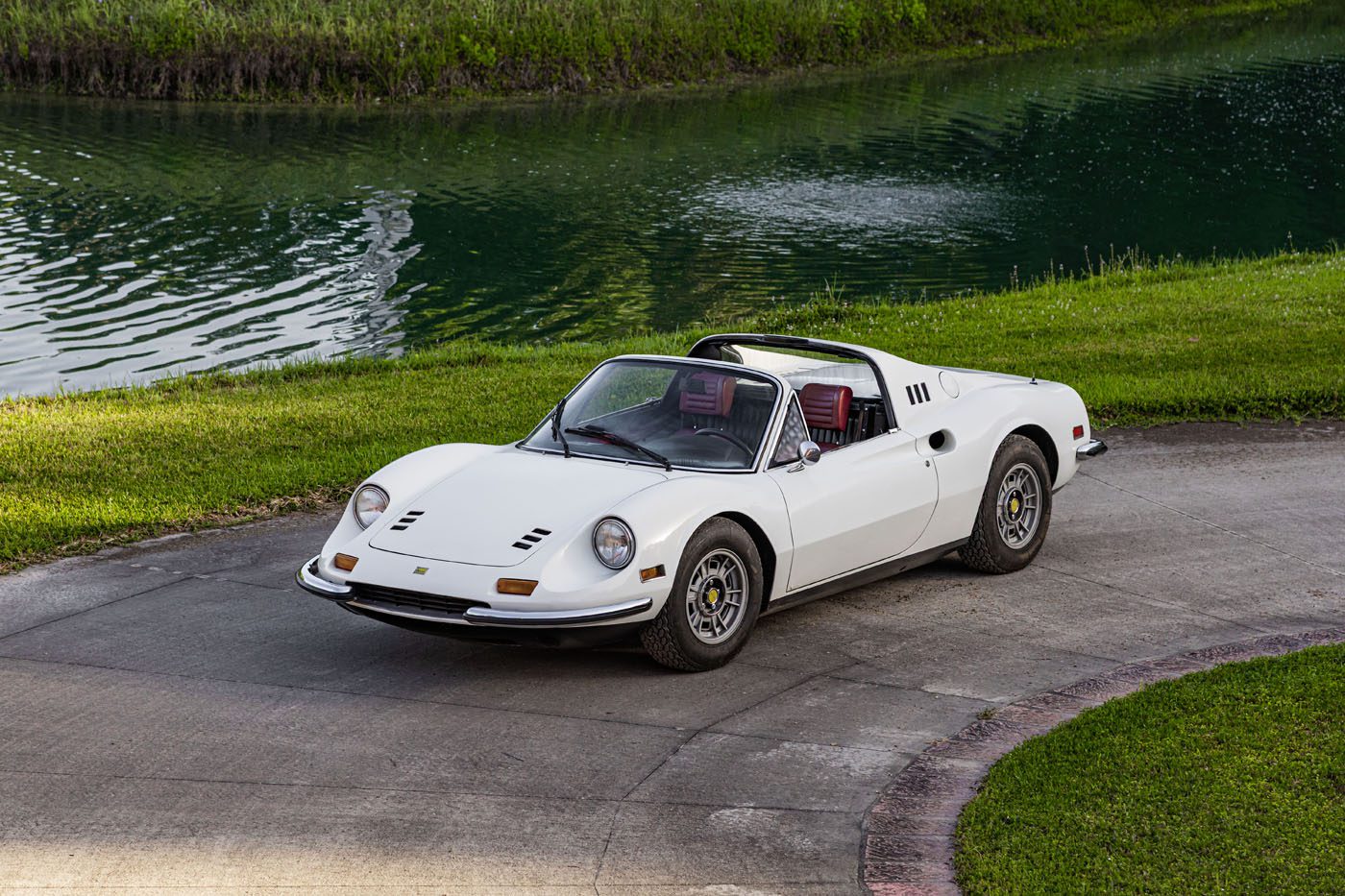 A classic white convertible sports car with a targa top is parked on a curved driveway near a grassy area and a pond, capturing the spirit of Ferrari Legends at Monterey ahead of Mecum’s 2025 Auction.