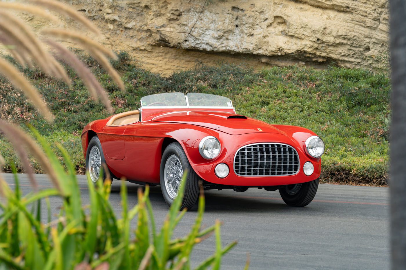 A classic red convertible sports car with chrome details is parked on a road, its vibrant finish echoing Ferrari Legends soon to be seen at Mecum’s 2025 Auction in Monterey, surrounded by greenery and a rocky hillside in the background.