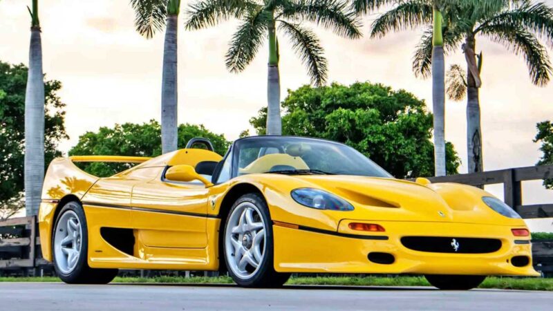 A yellow Ferrari F50 sports car, one of the 10 Most Expensive Cars To Sell At 2025 Monterey Car Week Auctions, parked outdoors near palm trees and viewed from a low front angle.