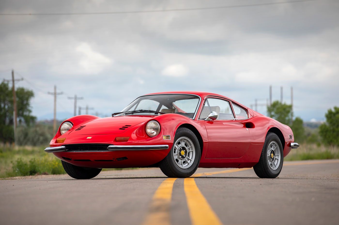 A red classic sports car, a true Ferrari Legend, is parked on a rural road under a cloudy sky, with trees and utility poles in the background—evoking the spirit of the Monterey auction.